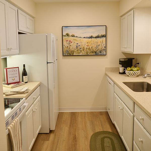 Kitchen area of a Putnam apartment at Pomperaug Woods showing white cabinets, a refrigerator, stove, sink, countertop appliances, a cookbook and bottle on the counter, and framed landscape artwork on the wall.