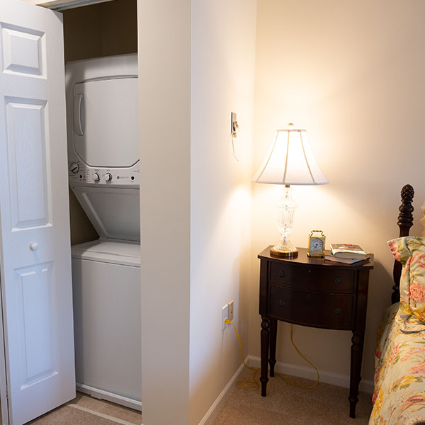 Interior corner of a Putnam apartment at Pomperaug Woods showing a stacked washer and dryer in an open closet next to a nightstand with lamp and part of a bed in a bedroom setting.